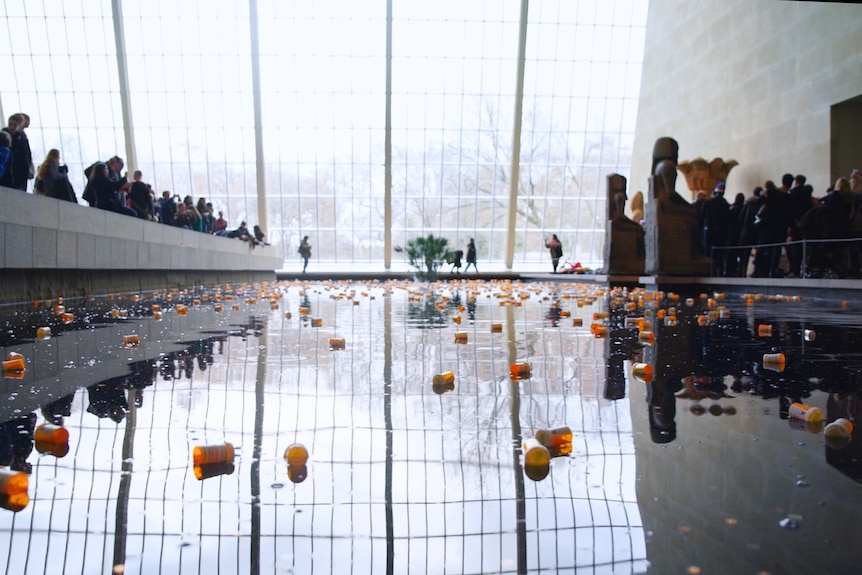 A moat around a temple in an art gallery is littered with orange prescription bottles, with protesters standing on each side