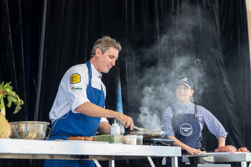 A chef wearing a blue apron cooking meat on a portable cooker.