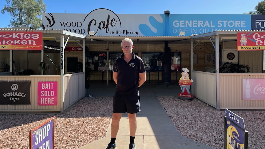 A man with short, white hair, wearing spectacles, a polo and shorts, stands in front of the Wood Wood cafe and general store.  