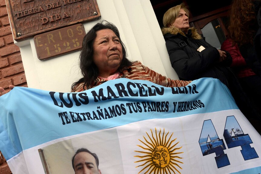 a woman holds a flag with the name of her son on it outside a building with another woman in the background