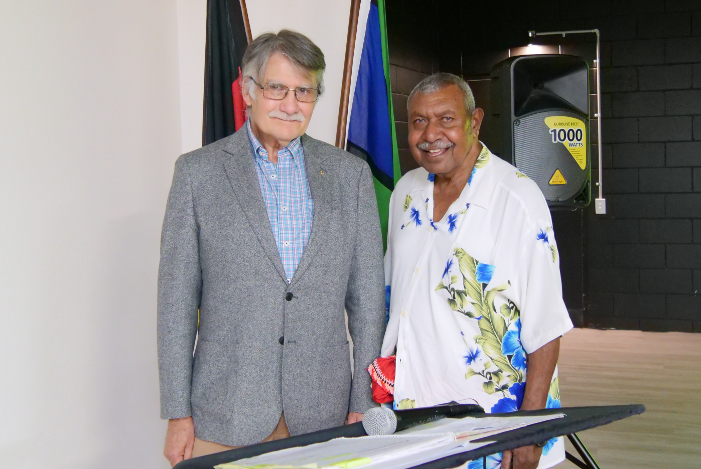 Two men standing next to each other next to a podium with Torres Strait and Aboriginal flags in the background
