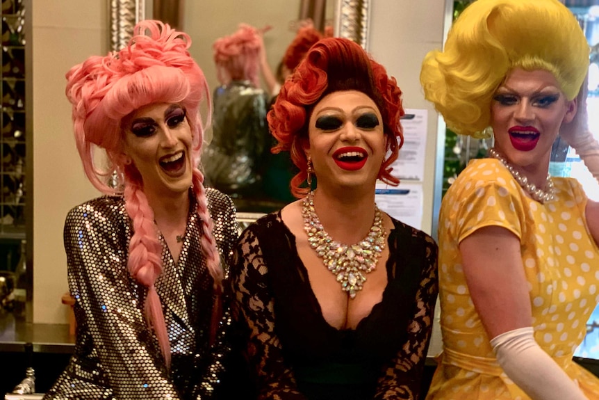 Three drag queens sit on a bar smiling