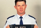 A fresh-faced police officer in blue shirt holds a black sign with his police number in white.