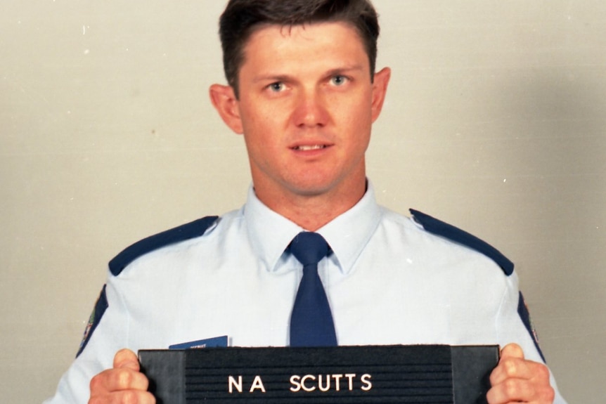 A fresh-faced police officer in blue shirt holds a black sign with his police number in white.