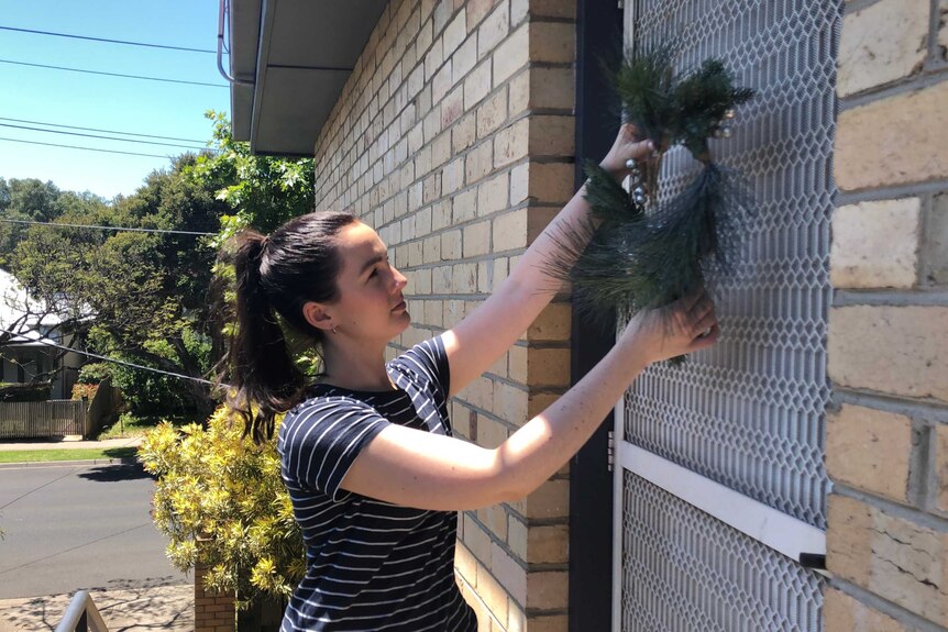 A woman wearing a black and white striped t-shirt hangs a Christmas wreath on her front door.
