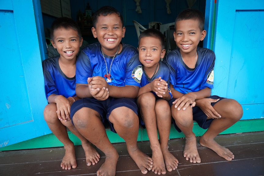Four smiling children in matching blue shirts sit squished together in a doorway. The walls are blue and floor is timber