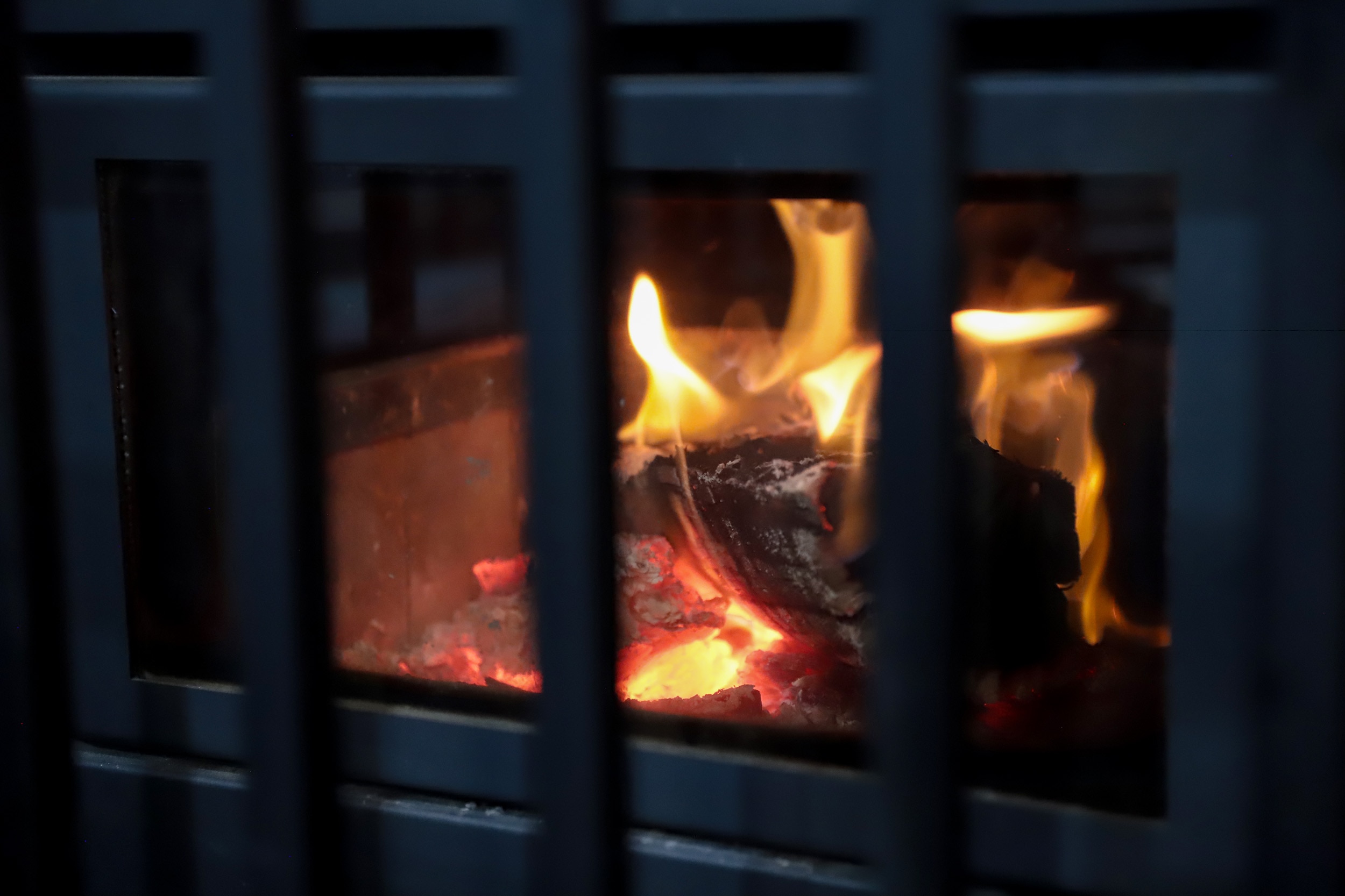 A burning log with embers and flames in a fireplace behind glass and metal grill bars.