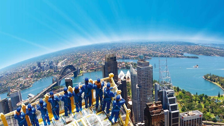 People stand on a glass platform high above the city, overlooking Sydney harbour.