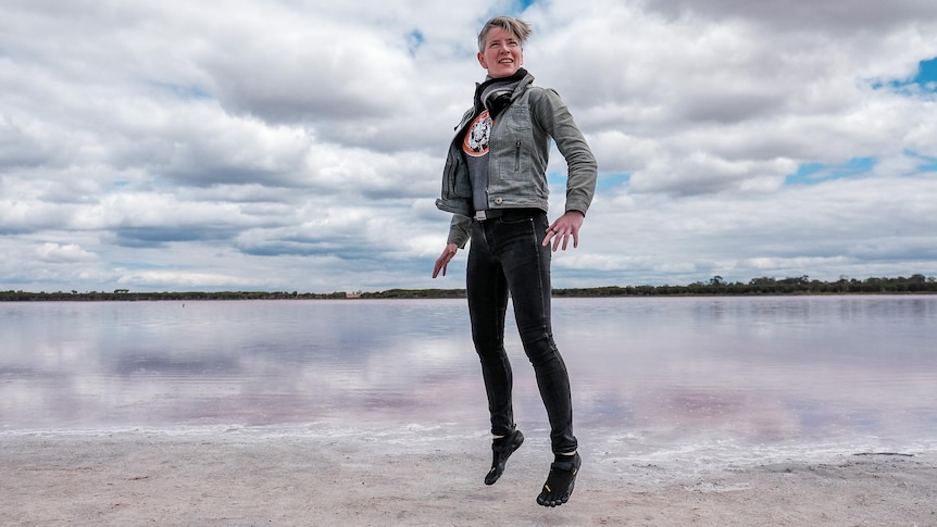 A woman jumps in the air in front of a pink lake and a cloudy sky.