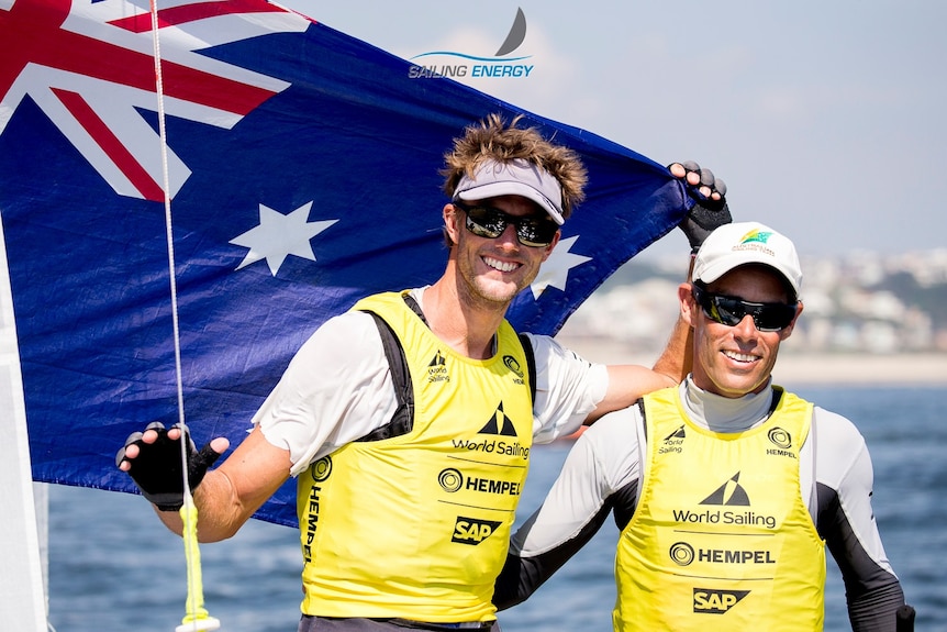 Two men wearing high-vis and sunglasses on a sailing boat with the Australian flag.