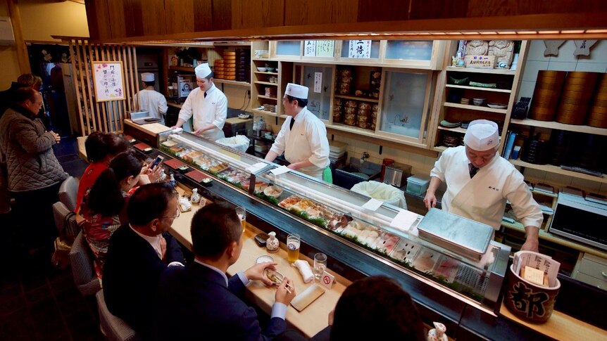 People sit in front of a sushi train as chefs prepare food on the other side.