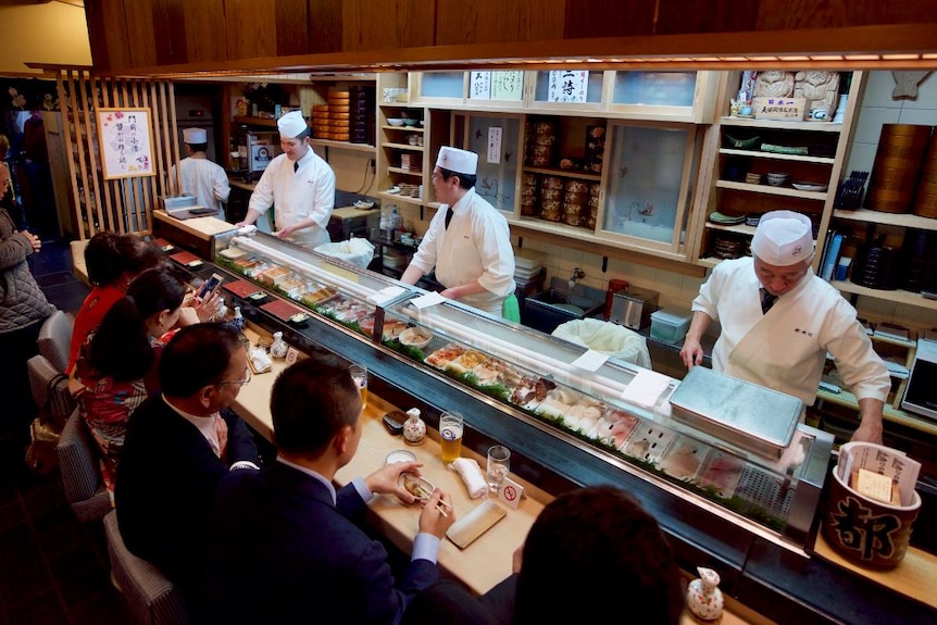 People sit in front of a sushi train as chefs prepare food on the other side.