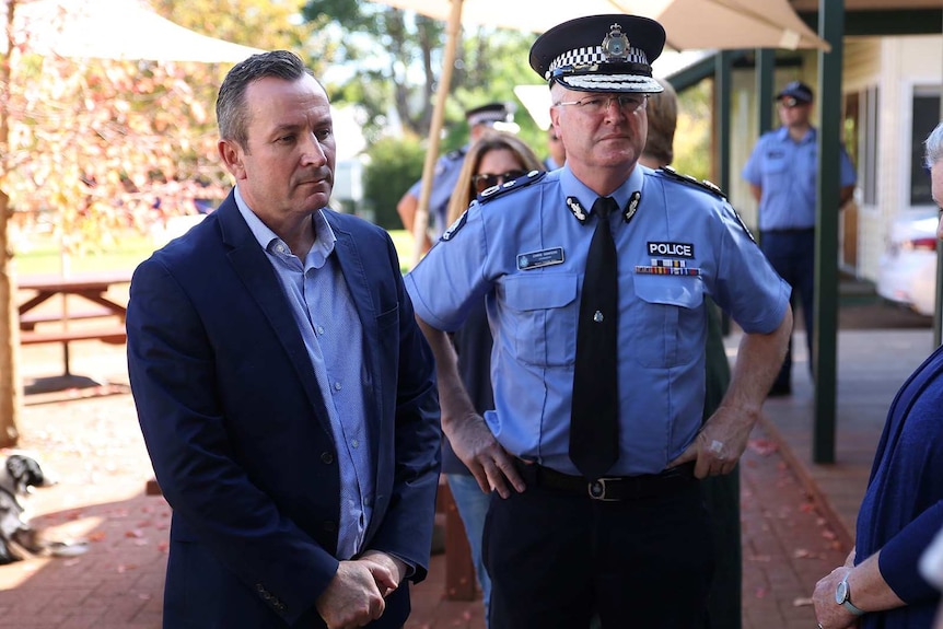 A man in a dark blue suit and a man in a police suit stand among a group of people outside.