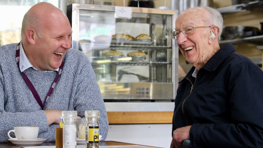An elderly man laughs heartily with a middle-aged man sitting next to him. There's one coffee on the table and pastires behind.