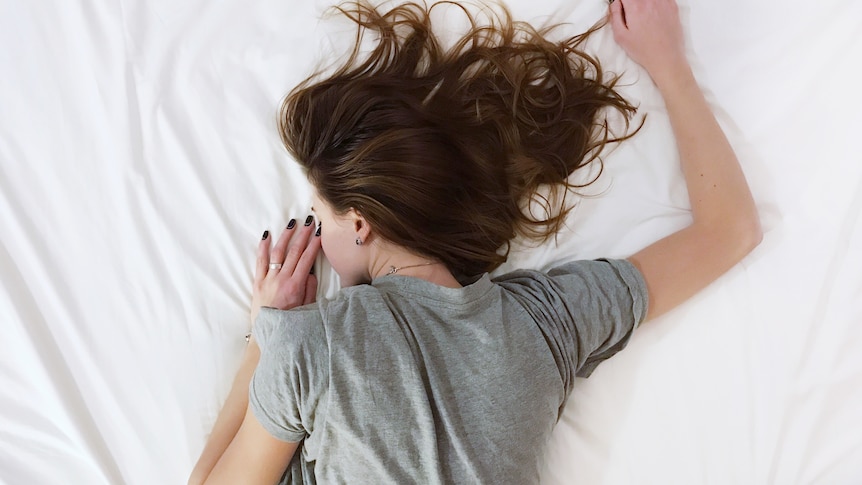 A woman laying face down in bed, with her hair sprawled over the sheets and her right arm above her head.