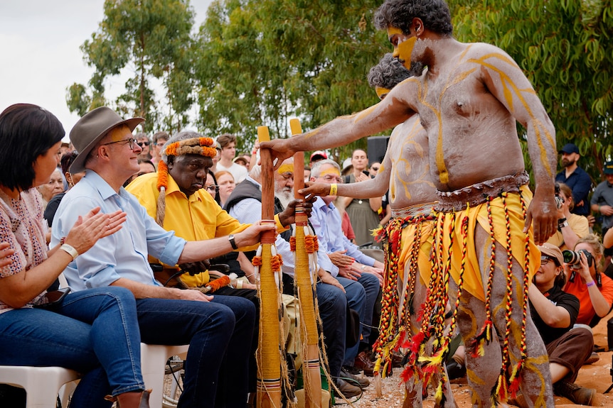 Two men, painted up, wearing traditional Aboriginal dress, present two men sitting down with cultural objects