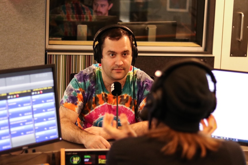 A dark-haired man in a tie-dyed shirt sits at a microphone inside a radio studio.
