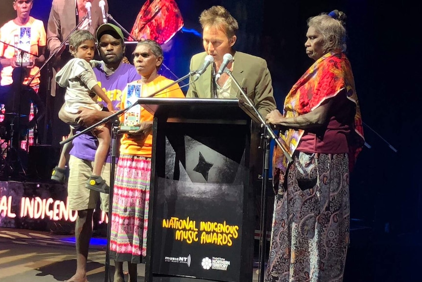Michael Hohnen and members of G. Yunupingu's family stand around a lectern, one speaks into a microphone