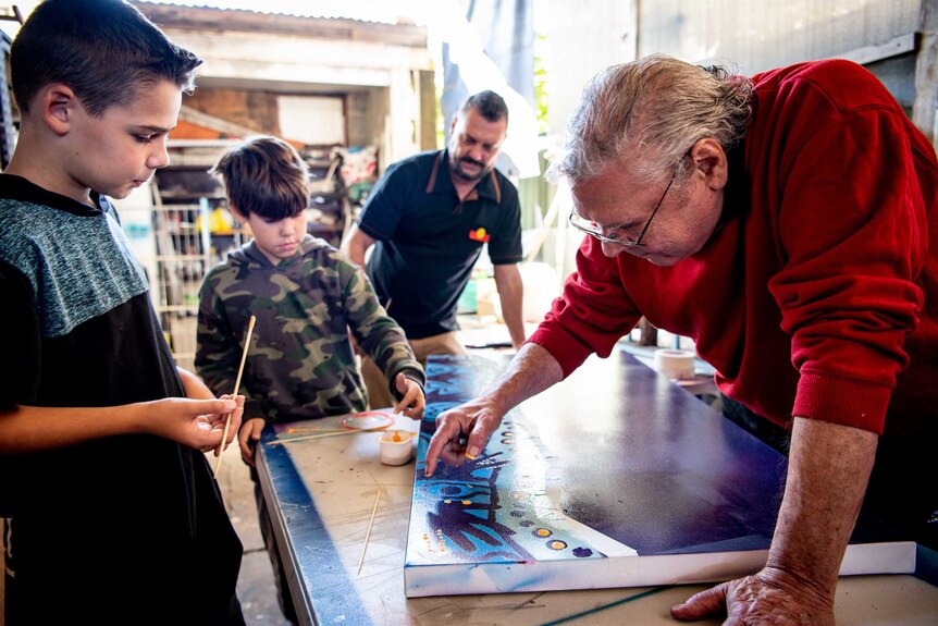 Danny leans over a painting on a table as his grandson watches on, with his son and other grandson in the background.