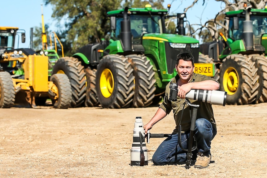 man sitting next to a big camera with tractors in the backgorund