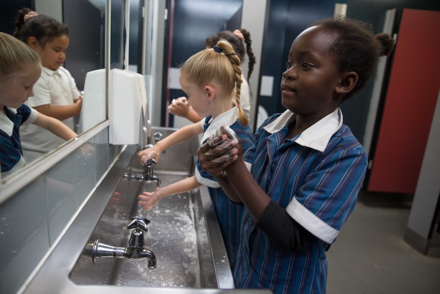 Three primary school students stand in front of the school bathroom mirror and wash their hands with soap