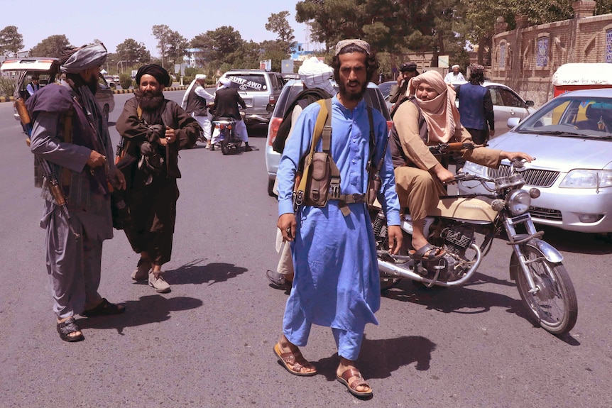 A young middle eastern man in a blue traditional suit walks in middle of street with woman on motorbike behind.