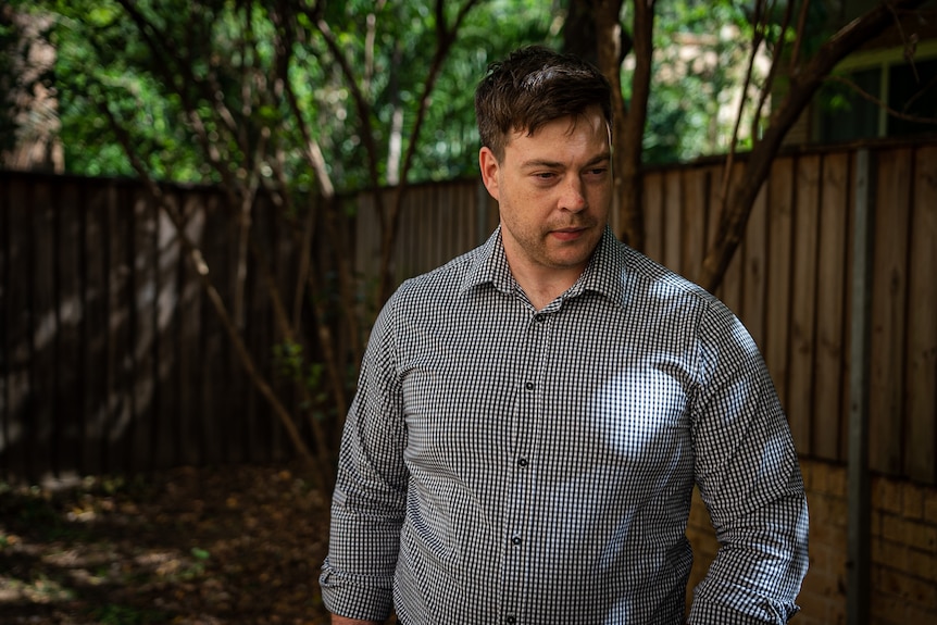 A man in a check shirt stands in his backyard looking down at the ground