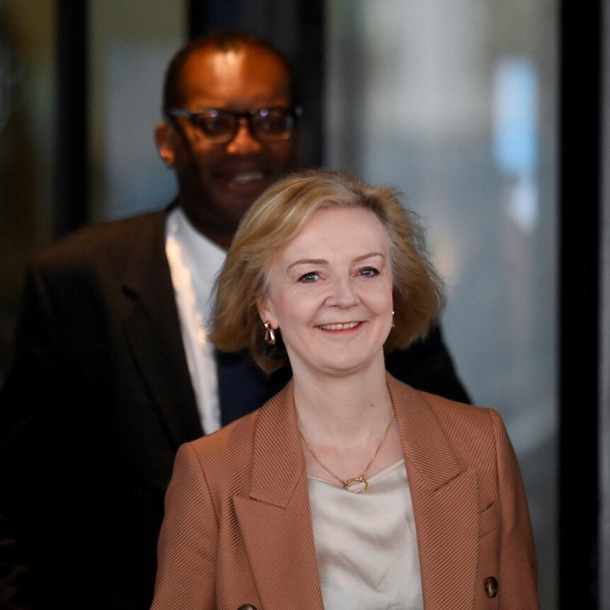 A short white middle-aged woman and a tall younger black man, both in suits, smile as they exit a glass door.