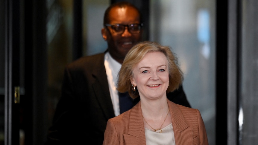 A short white middle-aged woman and a tall younger black man, both in suits, smile as they exit a glass door.