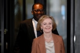 A short white middle-aged woman and a tall younger black man, both in suits, smile as they exit a glass door.