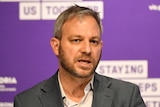Brett Sutton stands in front of a purple banner with Victorian Government logos on it.