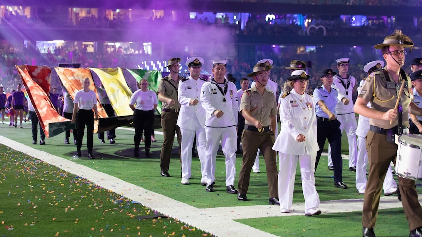 A parade of military officers marches in a stadium at night, carrying several banners that make up the colours of the rainbow.