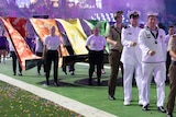 A parade of military officers marches in a stadium at night, carrying several banners that make up the colours of the rainbow.