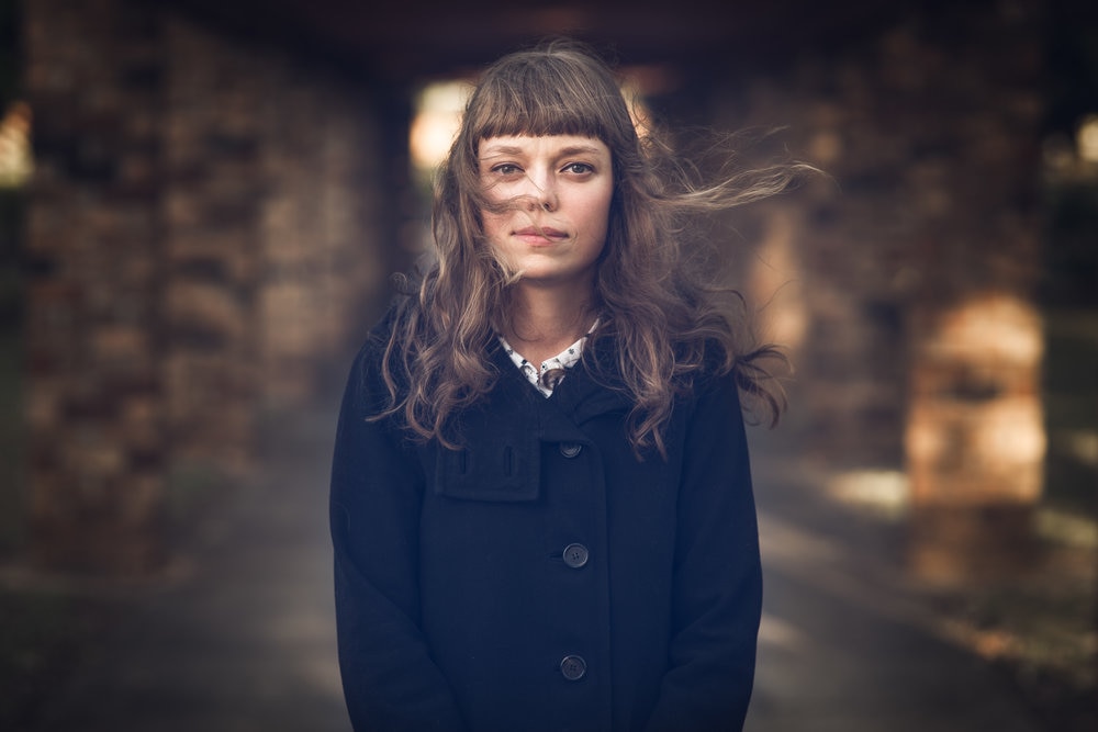 A young woman in a black coat with wavy hair blowing in her face, facing the camera and not smiling.
