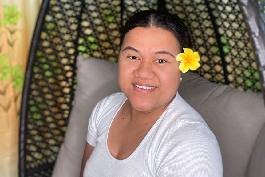 A woman with a white shirt and a yellow flower in her hair, smiles.