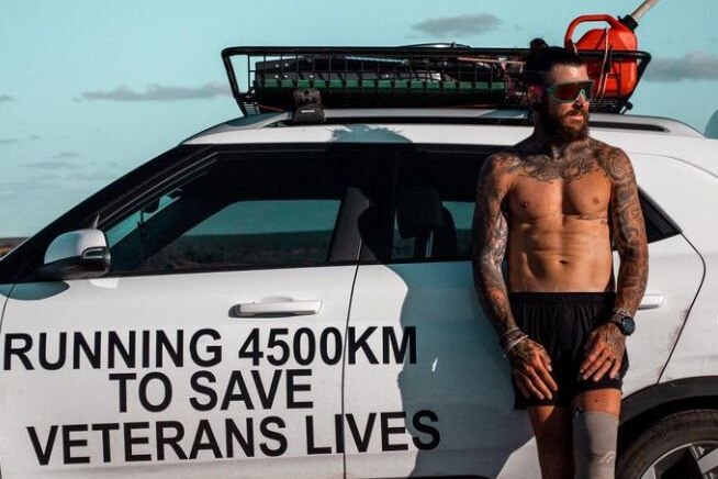 A shirtless man with sunglasses standing in front of a white car with black text on it looking at the sunset