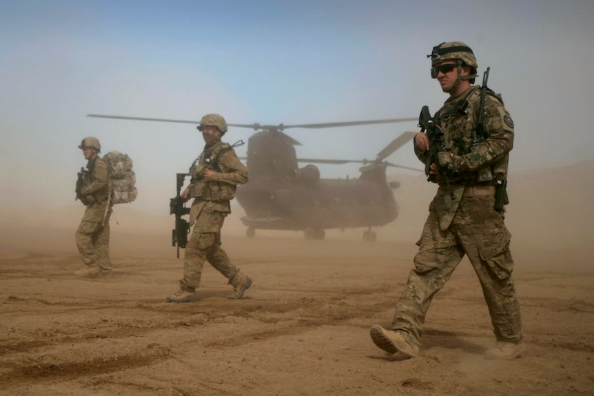 Three US soldiers in combat gear walking through a desert with a helicopter behind them 