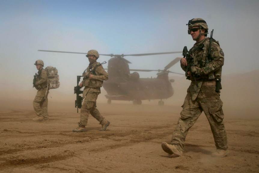 Trois soldats américains en tenue de combat marchant dans un désert avec un hélicoptère derrière eux 