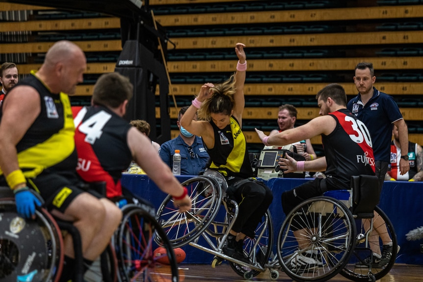 An action shot of Yasmina McGlone with arms stretched upwards with other players playing wheelchair AFL