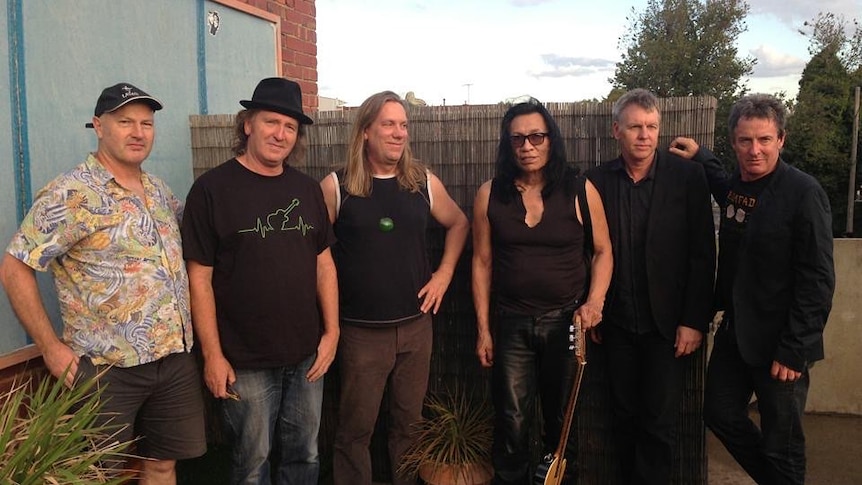 6 men standing in a suburban backyard. The man in the middle, Sixto Rodriguez, is holding an acoustic guitar.