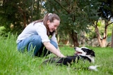 A teenager in jeans and a white shirt pats the belly of a smiling border collie lying in long grass.