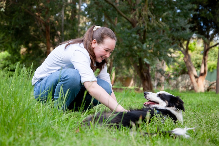 A teenage boy in jeans and a white shirt pats the stomach of a smiling border collie lying in tall grass.