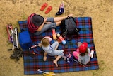A woman and two children sit on a rug for a family picnic. 