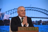 A middle-aged man with white hair puts his palms together in front of Sydney Harbour Bridge image