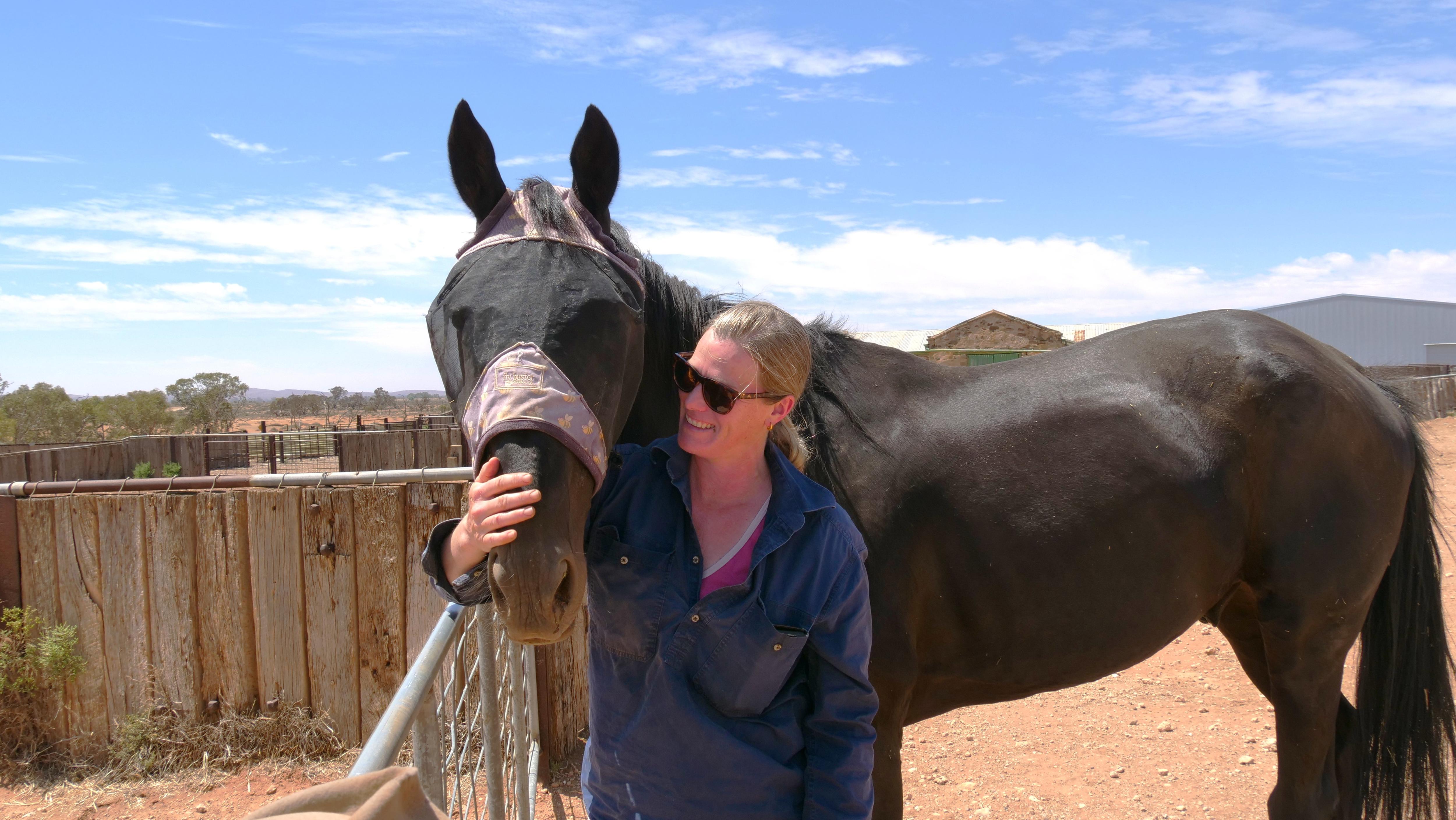 Una mujer con una camisa oscura sonríe y pone su brazo alrededor del cuello de un caballo oscuro en una propiedad rural