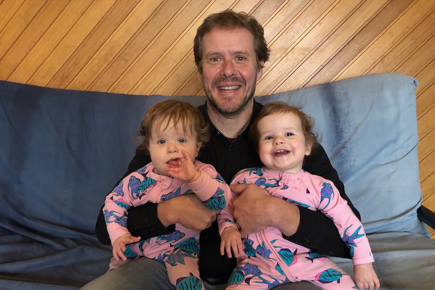 A dad sits on a blue futon couch with one baby twin in each arm, wearing matching pink jumpsuits