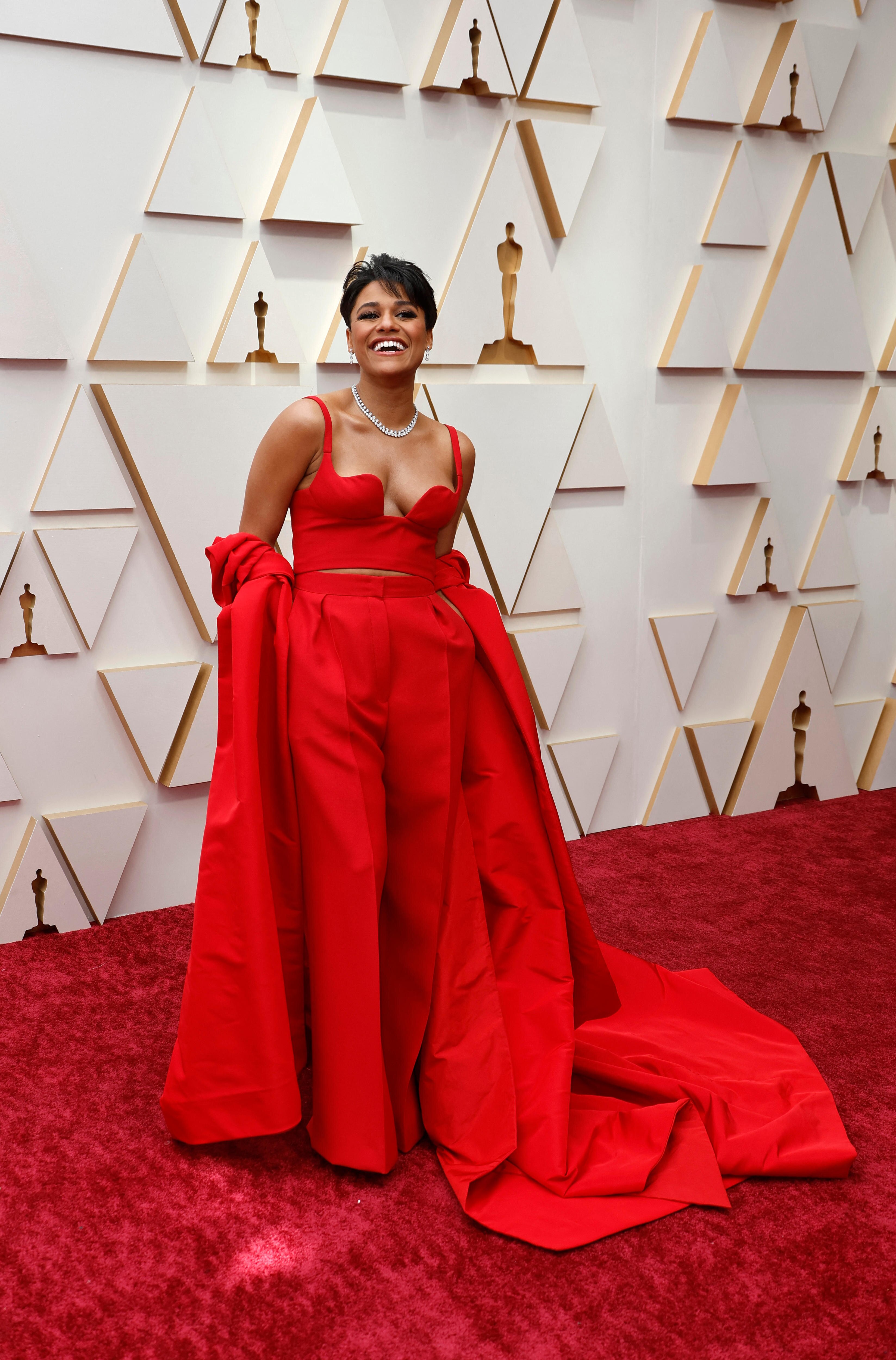 A woman wearing a red gown stands on a red carpet in front of a white media wall at the Oscars.