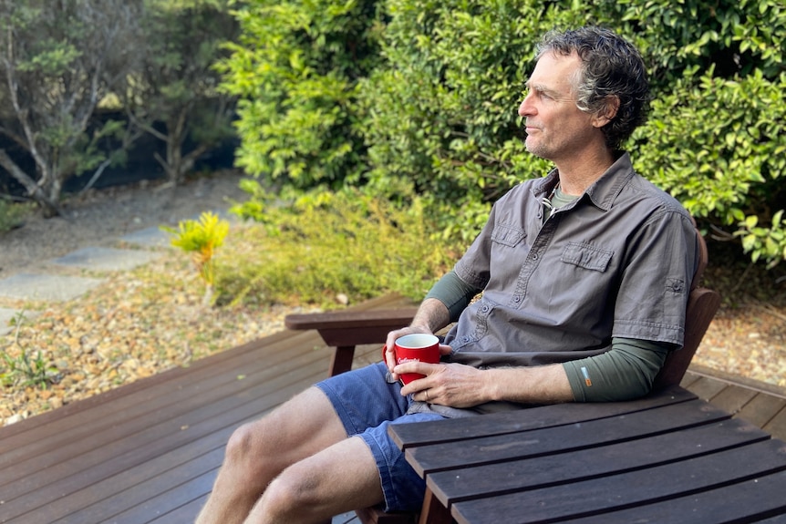 Man sitting on a woodern chair on an outside deck, holding a cup of coffee and looking out into his yard in the background