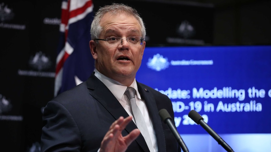 A man in a suit raises his right hand as he speaks into two microphones.