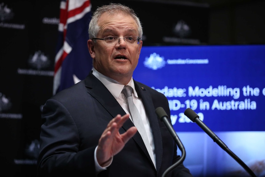 A man in a suit raises his right hand as he speaks into two microphones.